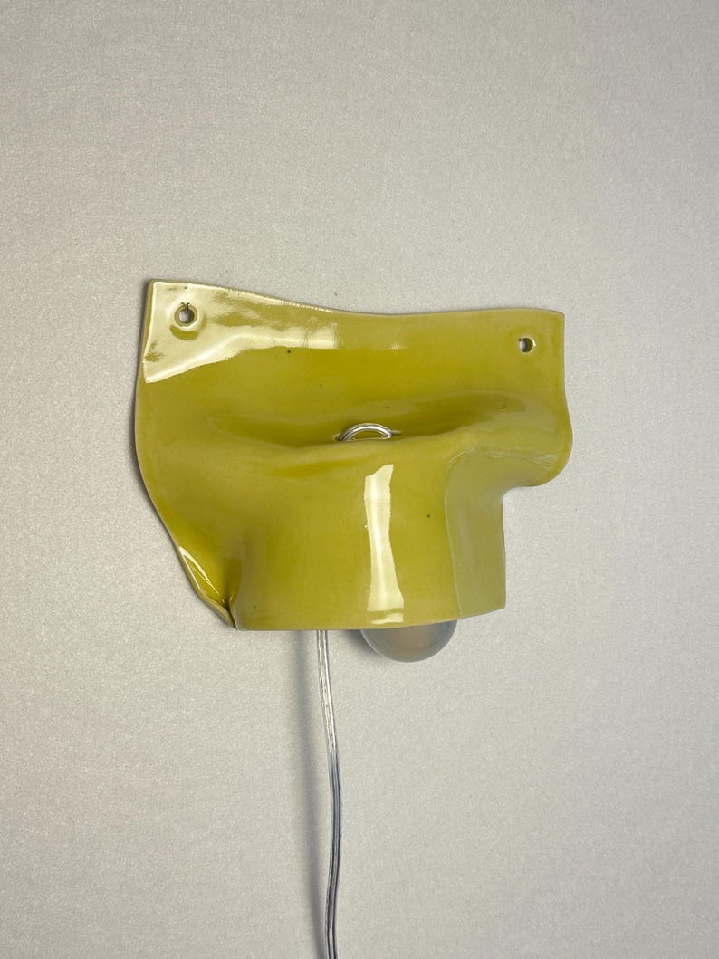 May include: A yellow wall-mounted light fixture with a bulb and a clear cord. The fixture has a unique, curved design with a small metal ring. The light is mounted on a neutral-colored wall.