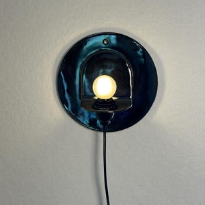 May include: A circular wall sconce featuring a glossy, deep teal ceramic backplate with a central arched cutout. A small, round, frosted light bulb is mounted within the cutout, emitting a warm glow. A thin black power cord extends downward.