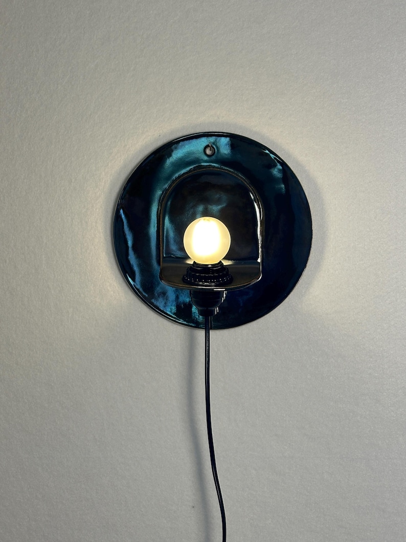 May include: A circular wall sconce featuring a glossy, deep teal ceramic backplate with a central arched cutout. A small, round, frosted light bulb is mounted within the cutout, emitting a warm glow. A thin black power cord extends downward.