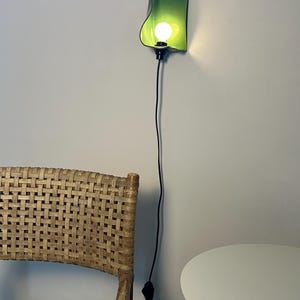 Pu&ograve; includere: Una lampada da parete con un paralume curvo in ceramica verde lime lucido e una lampadina accesa. L'apparecchio &egrave; collegato a un lungo e sottile cavo di alimentazione nero che pende verticalmente lungo la parete, terminando con un piccolo interruttore.