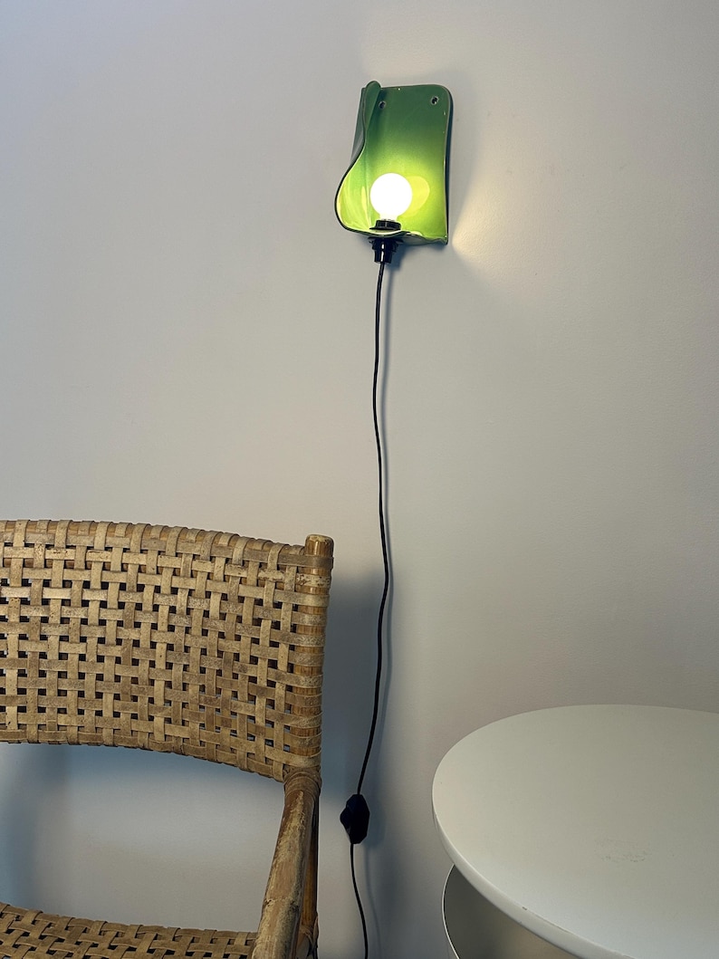 Pu&ograve; includere: Una lampada da parete con un paralume curvo in ceramica verde lime lucido e una lampadina accesa. L'apparecchio &egrave; collegato a un lungo e sottile cavo di alimentazione nero che pende verticalmente lungo la parete, terminando con un piccolo interruttore.