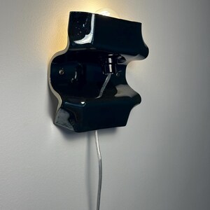 May include: A glossy black ceramic wall sconce with an organic, undulating shape. The fixture features a central light bulb socket emitting a warm glow from the top section and includes a thin, clear electrical cord extending downward from the base.