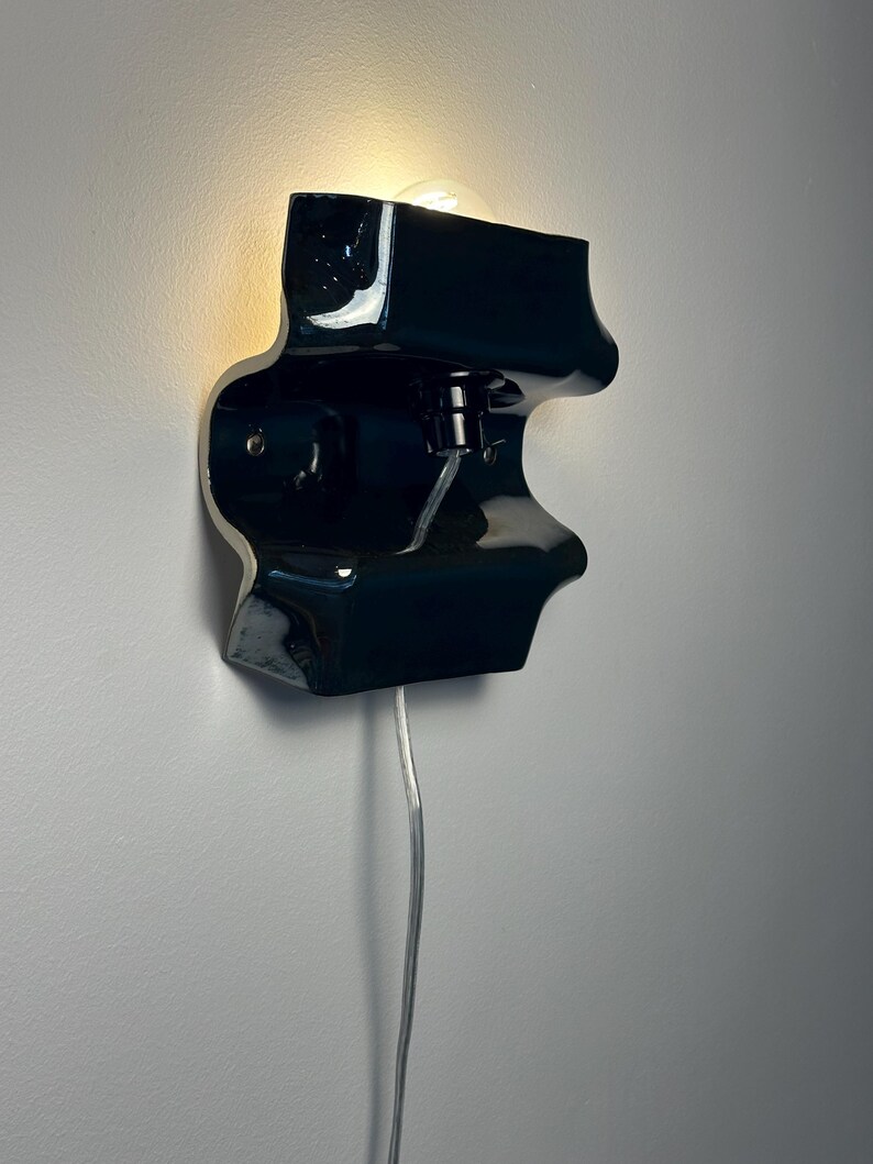 May include: A glossy black ceramic wall sconce with an organic, undulating shape. The fixture features a central light bulb socket emitting a warm glow from the top section and includes a thin, clear electrical cord extending downward from the base.