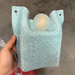 May include: A light blue wall sconce with a speckled finish and a smooth, rounded surface. The fixture features a central circular opening holding a spherical white light bulb and two small holes at the top corners for mounting to a wall surface.