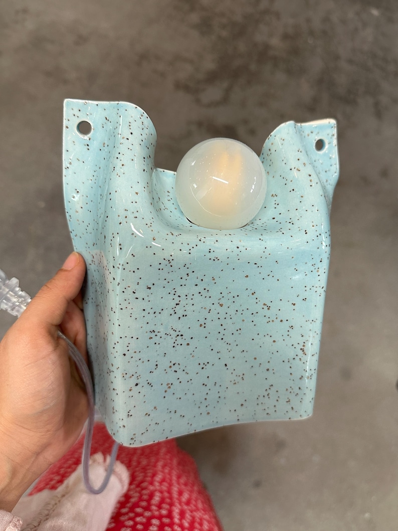 May include: A light blue wall sconce with a speckled finish and a smooth, rounded surface. The fixture features a central circular opening holding a spherical white light bulb and two small holes at the top corners for mounting to a wall surface.
