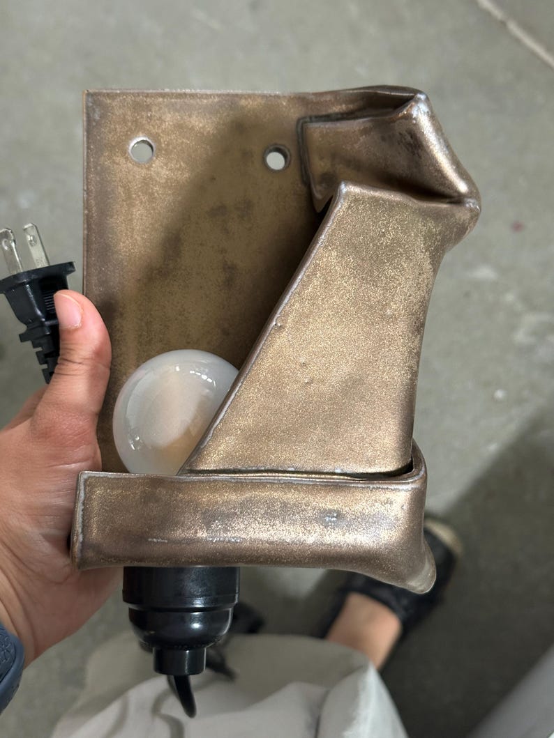 May include: A wall-mounted lamp fixture with a metallic bronze finish and a folded, sculptural design. The fixture includes a white spherical light bulb, a black electrical socket, and a black power cord with a standard two-prong plug.