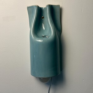 May include: A wall-mounted light fixture made of glossy, light blue ceramic. The piece features a sculptural, folded design with a central opening for a light bulb at the bottom. A small circular hole is visible near the top for mounting purposes.