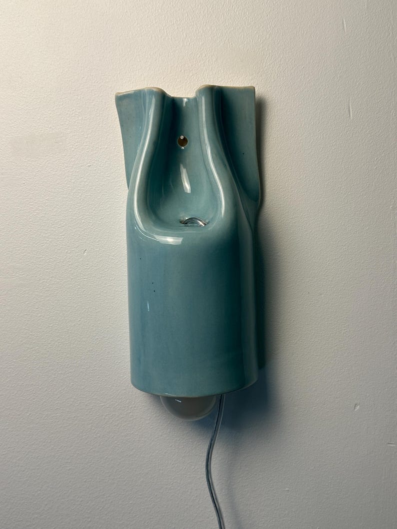 May include: A wall-mounted light fixture made of glossy, light blue ceramic. The piece features a sculptural, folded design with a central opening for a light bulb at the bottom. A small circular hole is visible near the top for mounting purposes.