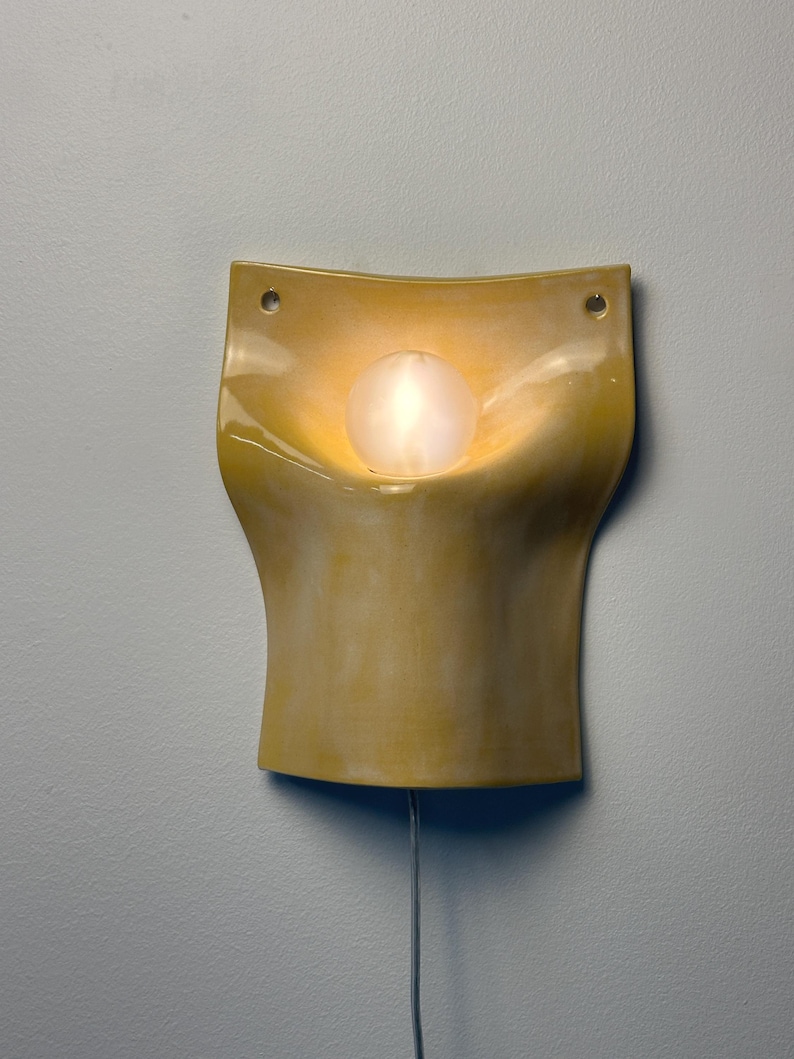 May include: A wall-mounted ceramic sconce with a glossy yellow finish and a curved, concave shape. The fixture features two circular mounting holes at the top corners and a central, glowing white spherical light bulb extending from the middle.