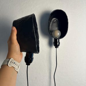 K&ouml;nnte beinhalten: Zwei schwarze Wandleuchten aus Keramik mit unregelm&auml;&szlig;igen, handgeformten Kanten. Jede Leuchte verf&uuml;gt &uuml;ber eine schwarze Fassung und ein schwarzes Kabel, wobei eine Leuchte mit einer runden wei&szlig;en Gl&uuml;hbirne ausgestattet ist. Die Oberfl&auml;che ist matt.