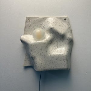 May include: A wall-mounted ceramic light fixture with an irregular, undulating surface featuring a white glaze with black speckles. The piece includes a single round, frosted glass bulb set into a recessed area and two small holes at the top.