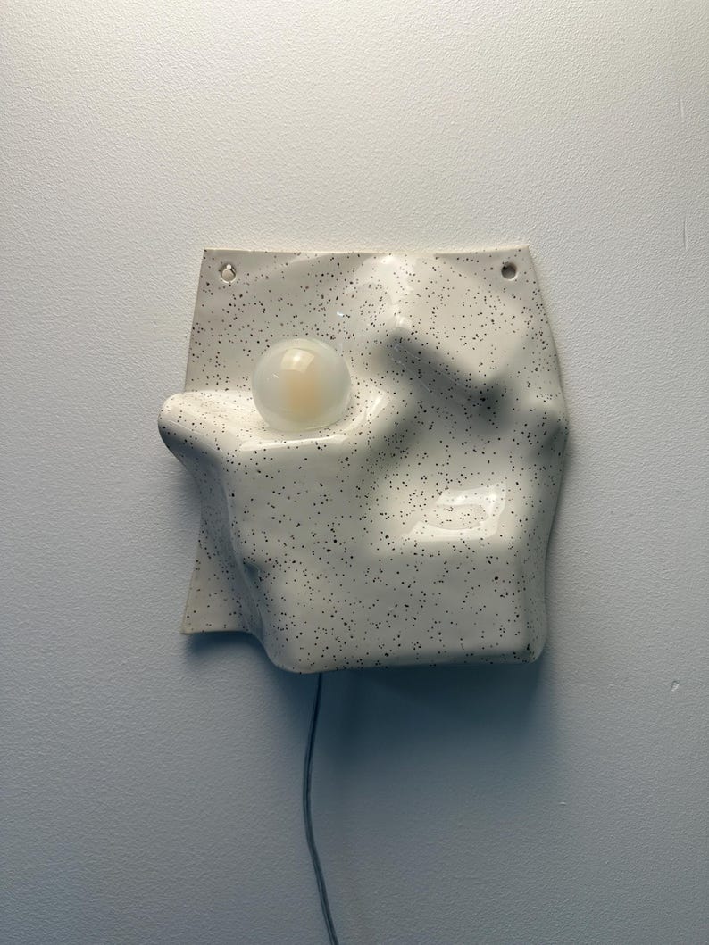 May include: A wall-mounted ceramic light fixture with an irregular, undulating surface featuring a white glaze with black speckles. The piece includes a single round, frosted glass bulb set into a recessed area and two small holes at the top.