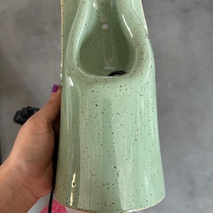 May include: A light green ceramic wall sconce with a speckled finish. The fixture features a tall, tapered, cylindrical body with a U-shaped cutout at the top and a small circular mounting hole. A black electrical cord extends from the base.