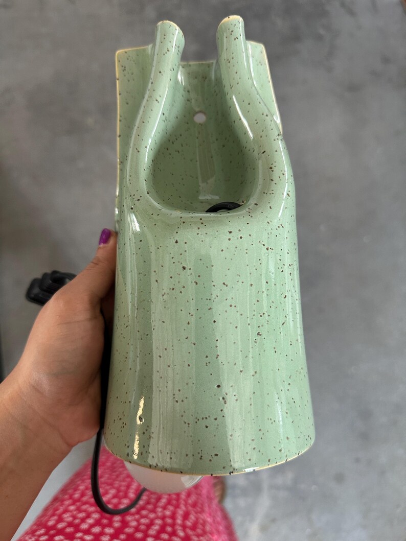 May include: A light green ceramic wall sconce with a speckled finish. The fixture features a tall, tapered, cylindrical body with a U-shaped cutout at the top and a small circular mounting hole. A black electrical cord extends from the base.