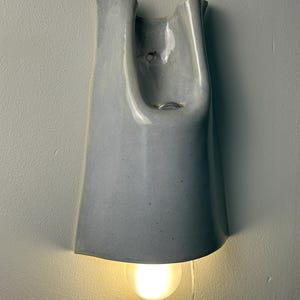K&ouml;nnte beinhalten: Eine Wandleuchte aus Keramik mit einer glatten, hellgrauen Glasur. Die Struktur weist ein hohes, konisch zulaufendes U-f&ouml;rmiges Design mit einer offenen Oberseite und einer abgerundeten Unterkante auf. Eine leuchtende Gl&uuml;hbirne ist an der Basis sichtbar.