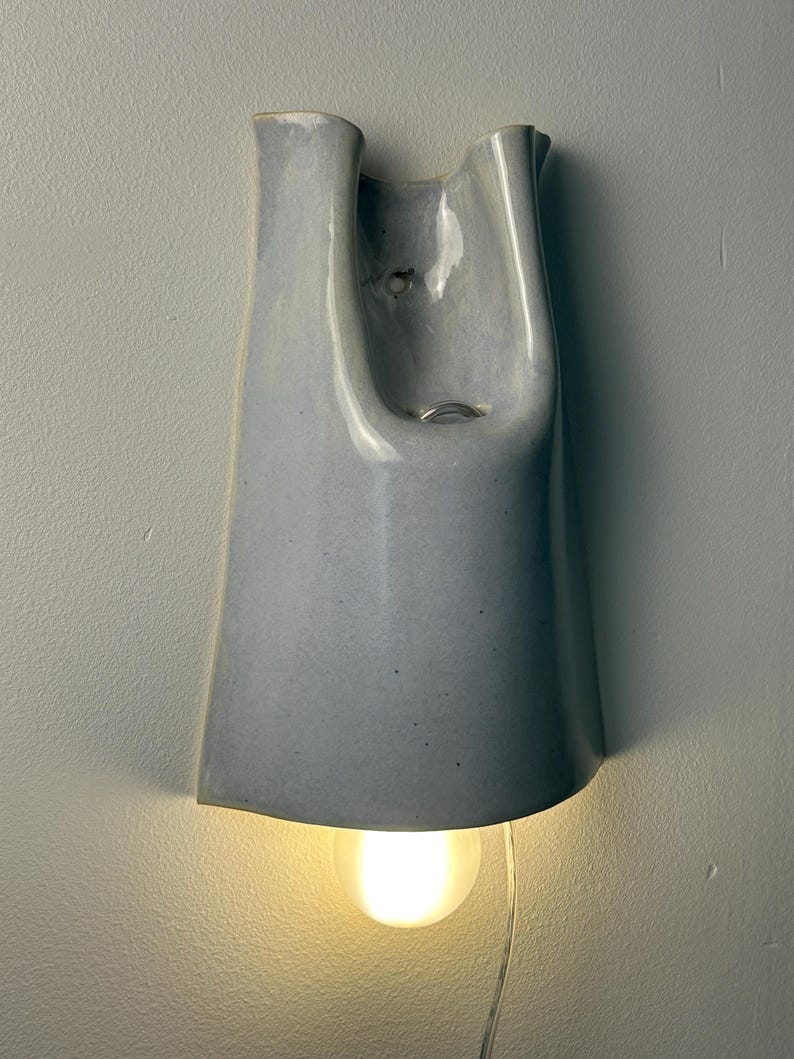 K&ouml;nnte beinhalten: Eine Wandleuchte aus Keramik mit einer glatten, hellgrauen Glasur. Die Struktur weist ein hohes, konisch zulaufendes U-f&ouml;rmiges Design mit einer offenen Oberseite und einer abgerundeten Unterkante auf. Eine leuchtende Gl&uuml;hbirne ist an der Basis sichtbar.