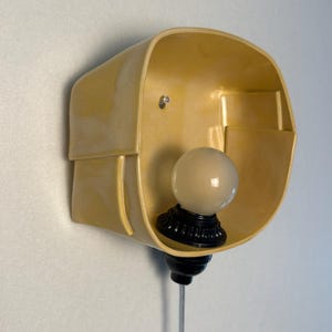 May include: A wall-mounted light fixture featuring a pale yellow, curved, bowl-shaped shade with a smooth, glossy finish. A black socket holds a single, round, frosted white light bulb at the base, with a thin, clear power cord extending downward.