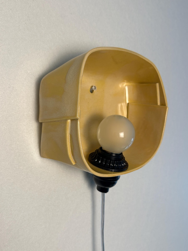 May include: A wall-mounted light fixture featuring a pale yellow, curved, bowl-shaped shade with a smooth, glossy finish. A black socket holds a single, round, frosted white light bulb at the base, with a thin, clear power cord extending downward.