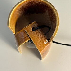 Puede incluir: Una l&aacute;mpara de mesa de cer&aacute;mica curva con un esmalte brillante de tonos &aacute;mbar y marr&oacute;n moteado. La estructura presenta una forma cil&iacute;ndrica abierta con un recorte central que sostiene un cable el&eacute;ctrico negro y una bombilla blanca visible.