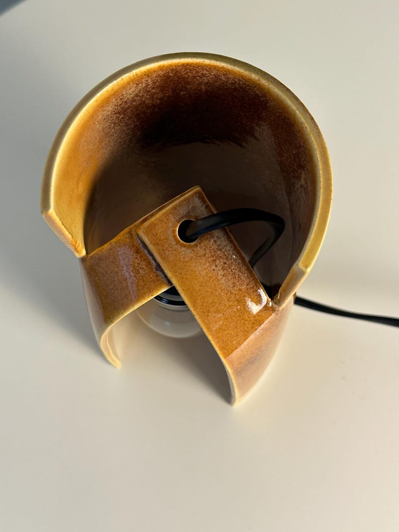 Puede incluir: Una l&aacute;mpara de mesa de cer&aacute;mica curva con un esmalte brillante de tonos &aacute;mbar y marr&oacute;n moteado. La estructura presenta una forma cil&iacute;ndrica abierta con un recorte central que sostiene un cable el&eacute;ctrico negro y una bombilla blanca visible.