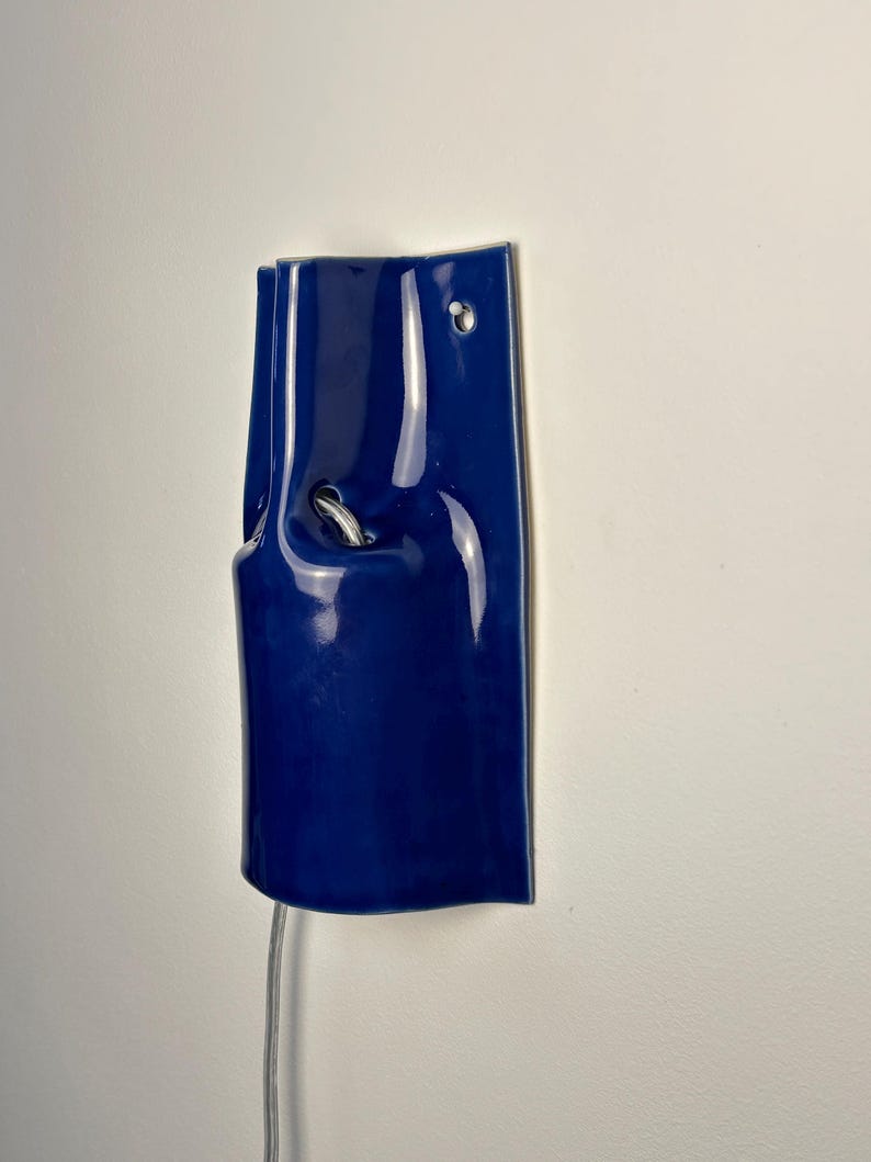 Puede incluir: Un aplique de pared de cer&aacute;mica azul oscuro brillante con forma rectangular curva. La l&aacute;mpara presenta una peque&ntilde;a abertura circular cerca de la parte superior para el montaje y un recorte central que revela un casquillo met&aacute;lico.