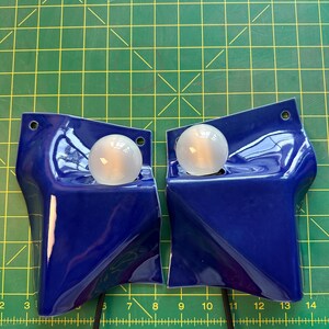 May include: Two glossy cobalt blue ceramic wall sconces with irregular, folded geometric shapes. Each fixture features a single round, frosted white light bulb set into a recessed area and a small circular mounting hole at the top corner.