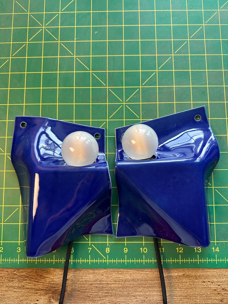 May include: Two glossy cobalt blue ceramic wall sconces with irregular, folded geometric shapes. Each fixture features a single round, frosted white light bulb set into a recessed area and a small circular mounting hole at the top corner.