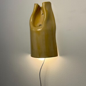 Pu&ograve; includere: Una lampada da parete in ceramica con una finitura lucida color giallo senape. La forma scultorea presenta una silhouette organica allungata con una sezione superiore cava e una lampadina che emette una luce calda dall'apertura inferiore.