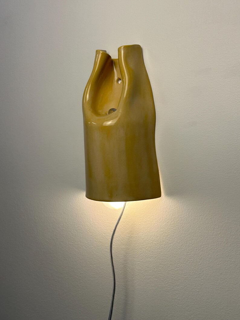 Pu&ograve; includere: Una lampada da parete in ceramica con una finitura lucida color giallo senape. La forma scultorea presenta una silhouette organica allungata con una sezione superiore cava e una lampadina che emette una luce calda dall'apertura inferiore.