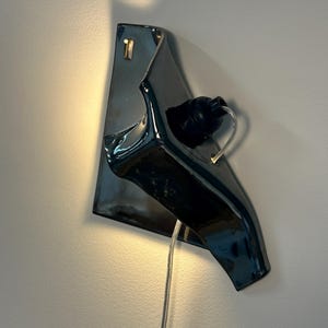 Peut inclure: Une applique murale compos&eacute;e d'une base en c&eacute;ramique gris anthracite brillant &agrave; la forme irr&eacute;guli&egrave;re et repli&eacute;e. Une douille noire est fix&eacute;e sur le devant, accueillant une ampoule transparente, avec un fin cordon d'alimentation transparent.