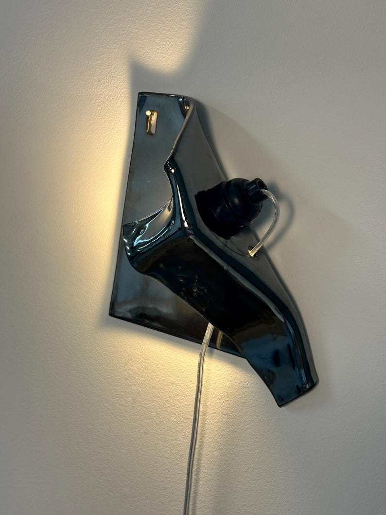 Peut inclure: Une applique murale compos&eacute;e d'une base en c&eacute;ramique gris anthracite brillant &agrave; la forme irr&eacute;guli&egrave;re et repli&eacute;e. Une douille noire est fix&eacute;e sur le devant, accueillant une ampoule transparente, avec un fin cordon d'alimentation transparent.