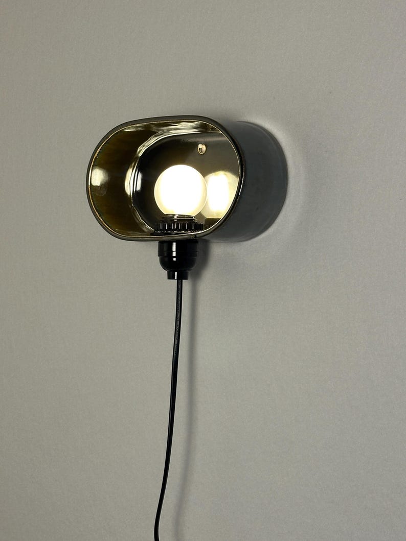 K&ouml;nnte beinhalten: Eine moderne Wandleuchte mit dunkelgrauer Au&szlig;enseite und goldfarbenem Innenraum. Die Leuchte hat eine ovale Form mit einer Gl&uuml;hbirne im Inneren. Ein schwarzes Kabel erstreckt sich von der Basis.