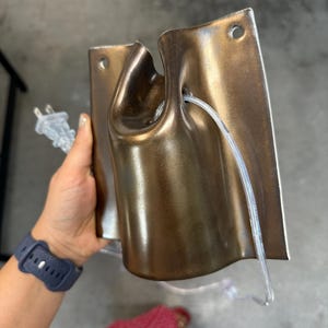 May include: A metallic bronze-colored wall-mounted light fixture with a curved, cylindrical center section and a flat rectangular backplate featuring two circular mounting holes. A clear electrical cord with a two-prong plug is threaded through.
