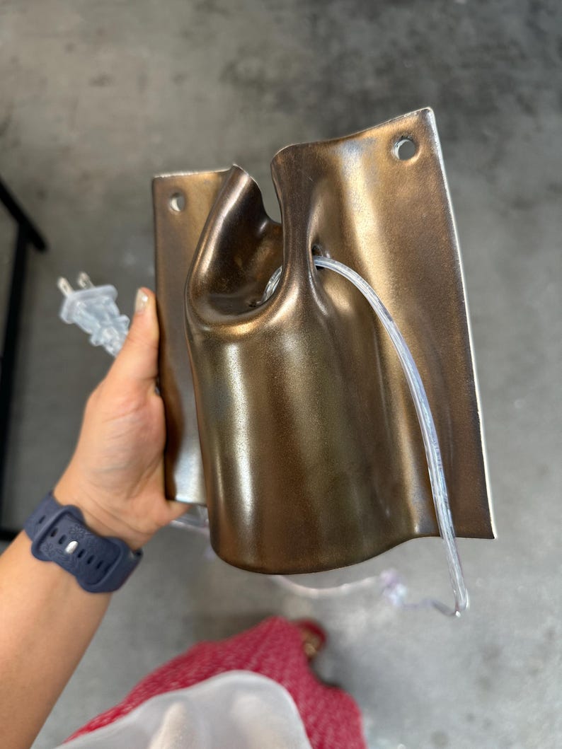 May include: A metallic bronze-colored wall-mounted light fixture with a curved, cylindrical center section and a flat rectangular backplate featuring two circular mounting holes. A clear electrical cord with a two-prong plug is threaded through.