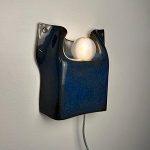 May include: A wall-mounted ceramic sconce featuring a deep blue, glossy glaze with a sculpted, organic shape. The fixture includes a central circular cutout holding a glowing, frosted white spherical light bulb and a thin power cord extending downward.