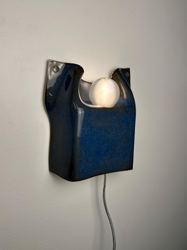 May include: A wall-mounted ceramic sconce featuring a deep blue, glossy glaze with a sculpted, organic shape. The fixture includes a central circular cutout holding a glowing, frosted white spherical light bulb and a thin power cord extending downward.