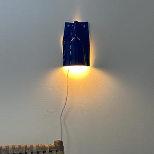 Puede incluir: Una l&aacute;mpara de pared de color azul oscuro con una pantalla cil&iacute;ndrica y una placa trasera plana. La l&aacute;mpara emite una luz c&aacute;lida dirigida hacia abajo que ilumina la pared. Un cable de alimentaci&oacute;n blanco cuelga verticalmente de la base.