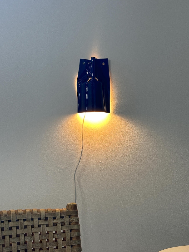 Puede incluir: Una l&aacute;mpara de pared de color azul oscuro con una pantalla cil&iacute;ndrica y una placa trasera plana. La l&aacute;mpara emite una luz c&aacute;lida dirigida hacia abajo que ilumina la pared. Un cable de alimentaci&oacute;n blanco cuelga verticalmente de la base.