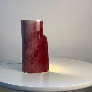 May include: A cylindrical ceramic table lamp with a glossy, deep red and burgundy gradient glaze. The lamp features an asymmetrical, stepped silhouette with a taller section and a shorter, rounded side. A warm light glows from the base.