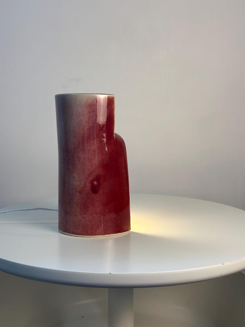 May include: A cylindrical ceramic table lamp with a glossy, deep red and burgundy gradient glaze. The lamp features an asymmetrical, stepped silhouette with a taller section and a shorter, rounded side. A warm light glows from the base.