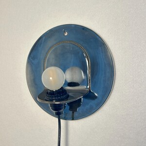 May include: A wall-mounted light fixture featuring a circular, glossy blue ceramic backplate with a metallic sheen. A black socket holds a single, round, frosted white light bulb. A black power cord extends downward from the base of the socket.