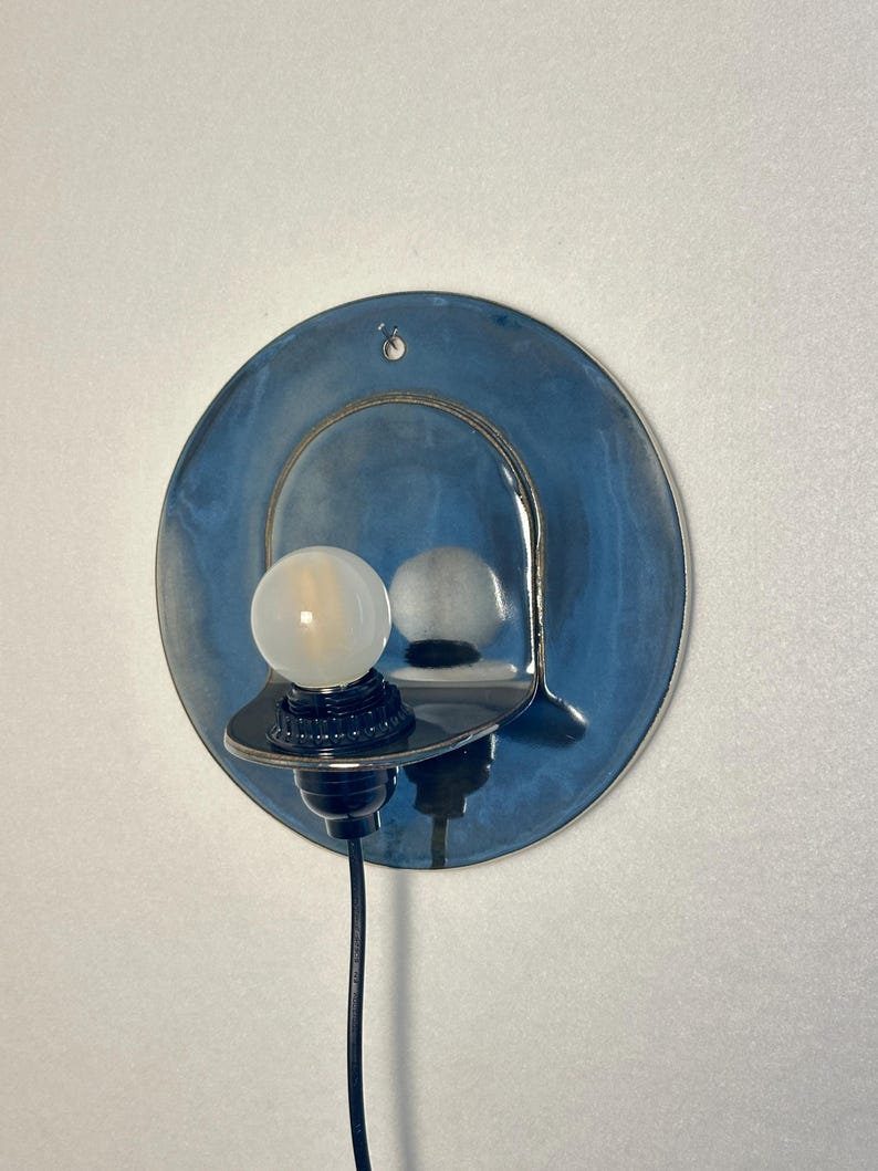 May include: A wall-mounted light fixture featuring a circular, glossy blue ceramic backplate with a metallic sheen. A black socket holds a single, round, frosted white light bulb. A black power cord extends downward from the base of the socket.