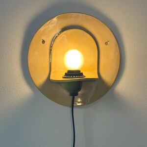Pu&ograve; includere: Un'applique circolare in ceramica gialla con una sporgenza curva a forma di cappuccio che ospita una lampadina accesa. L'apparecchio include un cavo di alimentazione nero che scende verso il basso e due piccoli fori di montaggio sulla piastra.