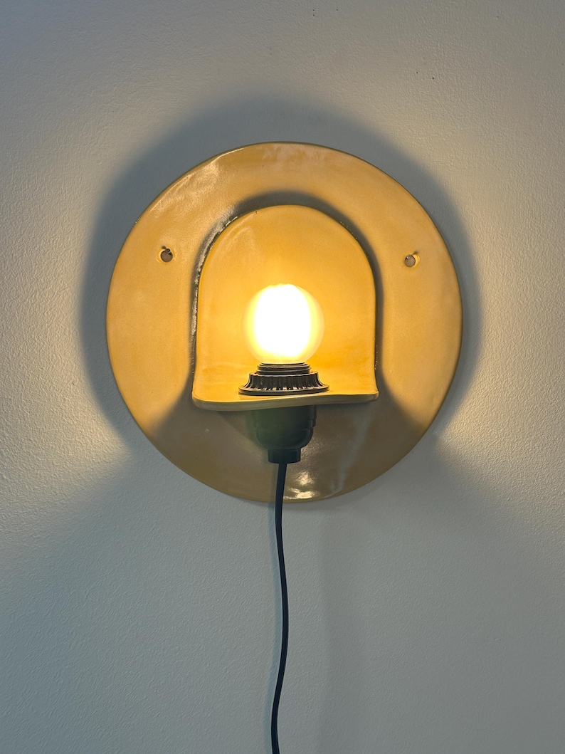 Pu&ograve; includere: Un'applique circolare in ceramica gialla con una sporgenza curva a forma di cappuccio che ospita una lampadina accesa. L'apparecchio include un cavo di alimentazione nero che scende verso il basso e due piccoli fori di montaggio sulla piastra.