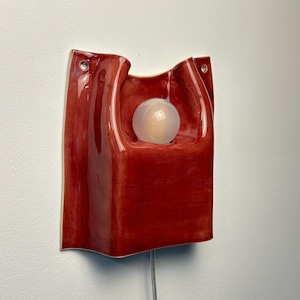 Puede incluir: Un aplique de pared con un cuerpo de cer&aacute;mica de color rojo intenso brillante y forma org&aacute;nica ondulada. La l&aacute;mpara incluye un recorte circular central que sostiene una bombilla esf&eacute;rica blanca esmerilada y dos orificios para el montaje.