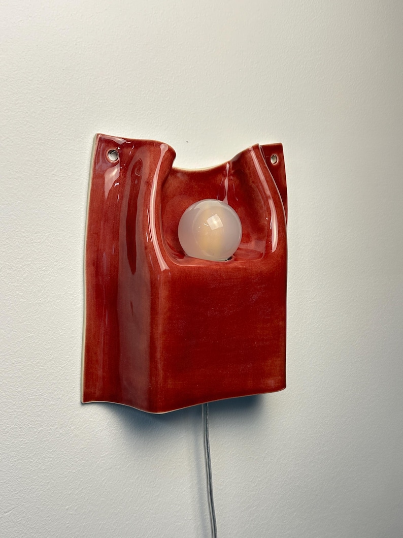 Puede incluir: Un aplique de pared con un cuerpo de cer&aacute;mica de color rojo intenso brillante y forma org&aacute;nica ondulada. La l&aacute;mpara incluye un recorte circular central que sostiene una bombilla esf&eacute;rica blanca esmerilada y dos orificios para el montaje.
