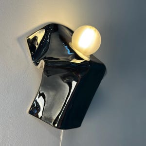 May include: A wall-mounted light fixture featuring a glossy, black, irregularly shaped ceramic base with a fluid, melted appearance. A single, spherical, frosted white glass bulb is positioned on the upper section, emitting a warm, soft glow.