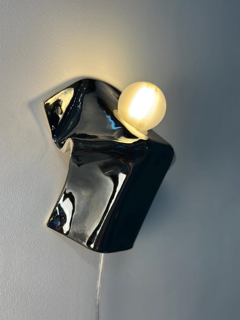 May include: A wall-mounted light fixture featuring a glossy, black, irregularly shaped ceramic base with a fluid, melted appearance. A single, spherical, frosted white glass bulb is positioned on the upper section, emitting a warm, soft glow.