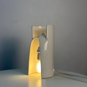 May include: A white ceramic table lamp with a curved, cylindrical structure that is open on one side. The lamp features a single glowing light bulb housed within the ceramic frame and includes a thin white power cord extending from the side.