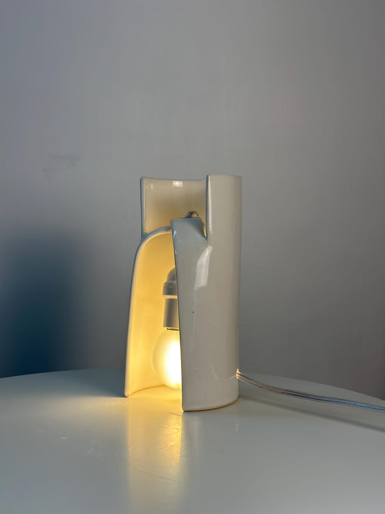 May include: A white ceramic table lamp with a curved, cylindrical structure that is open on one side. The lamp features a single glowing light bulb housed within the ceramic frame and includes a thin white power cord extending from the side.